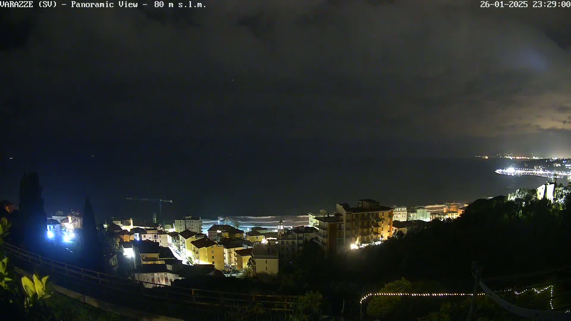 A nighttime panoramic view shows a coastal town with lights illuminating buildings overlooking a dark ocean under a cloudy sky.