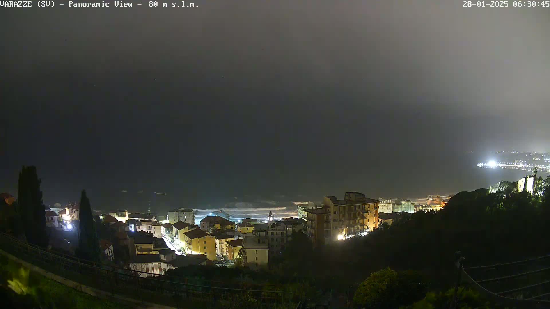 A nighttime, foggy view of a coastal town with its lights illuminated against a dark sea with some waves.
