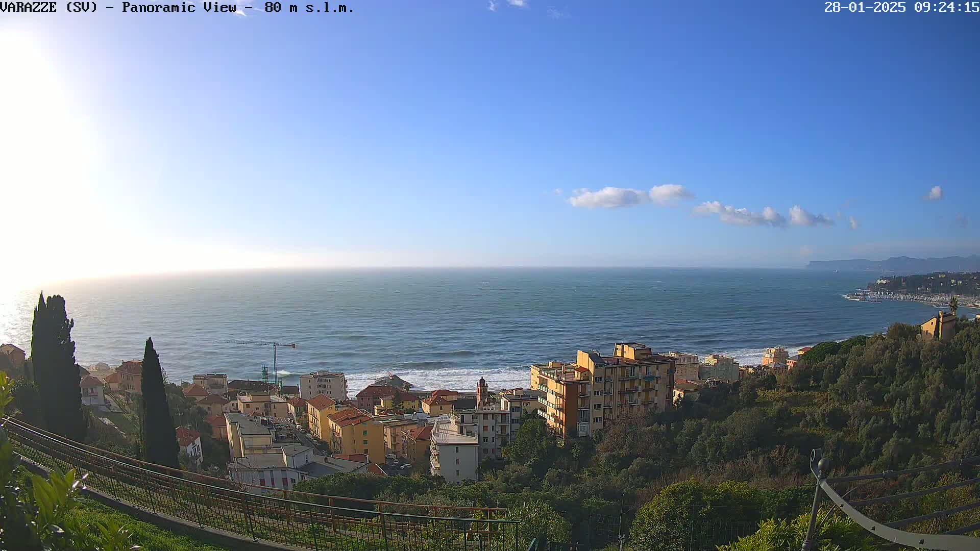 A coastal town nestled on a hillside overlooks a calm ocean under a mostly clear, blue sky with a few scattered clouds.