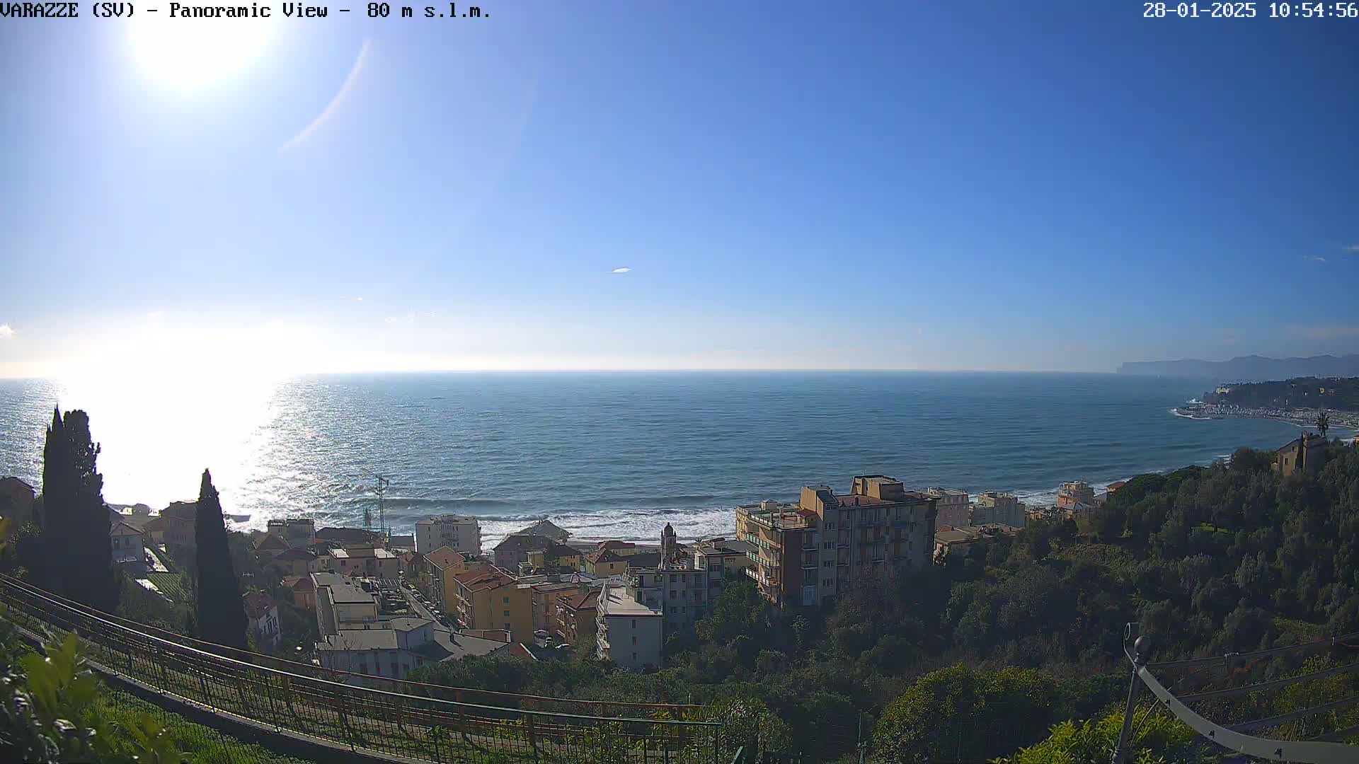 A panoramic view shows a town nestled on a hillside overlooking a calm sea under a clear blue sky.