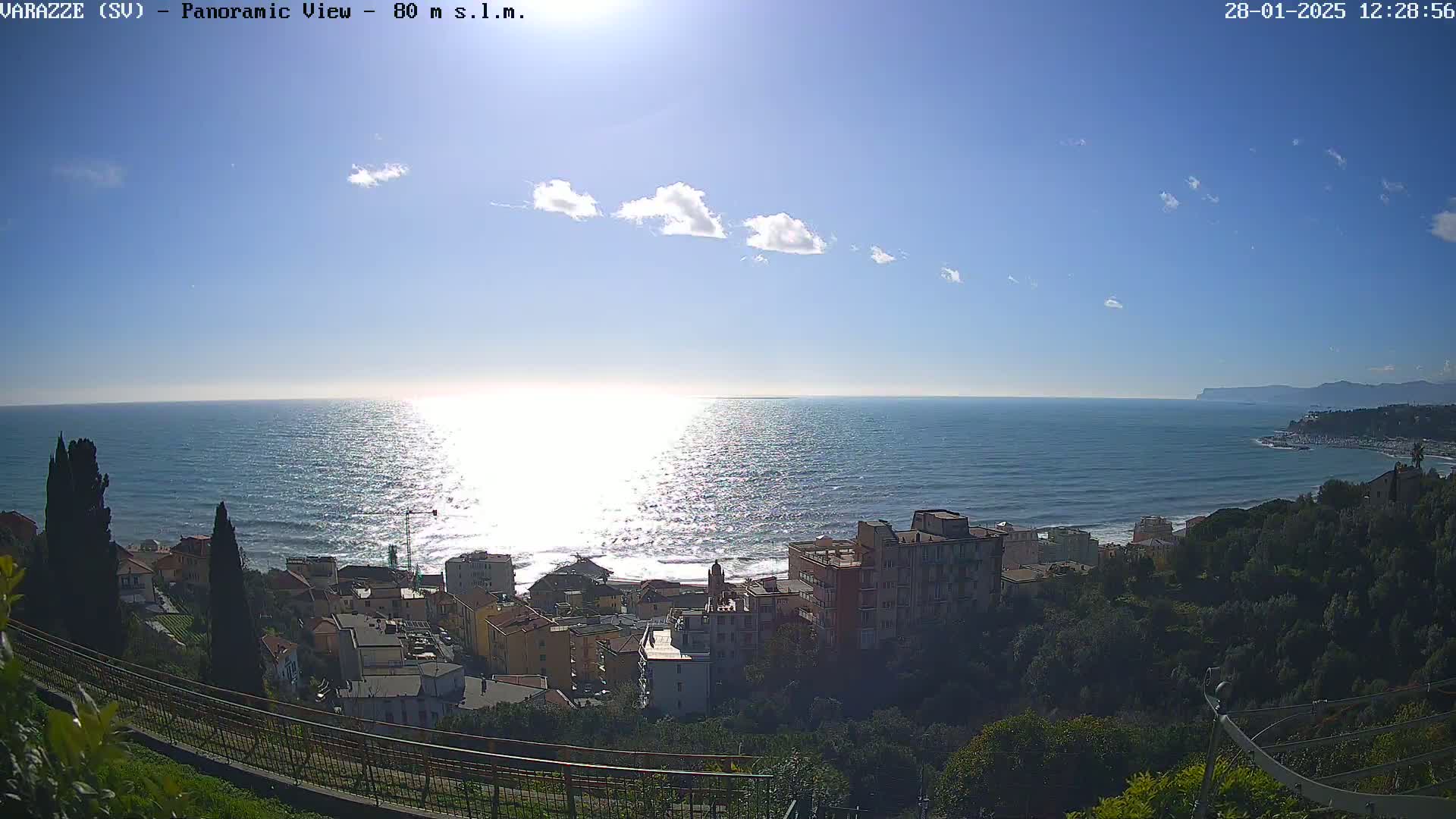 A coastal town nestled on a hillside overlooks a sparkling blue sea under a mostly sunny sky with a few scattered clouds.