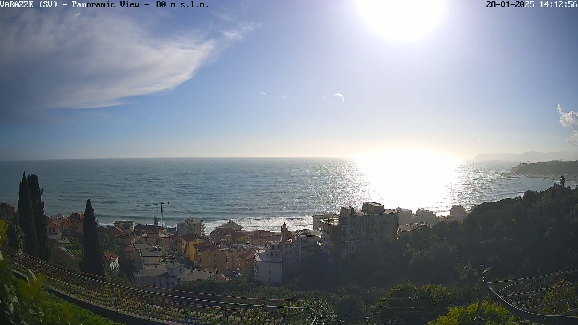 A coastal town nestled on a hillside overlooks a sparkling sea under a mostly sunny sky with a few scattered clouds.