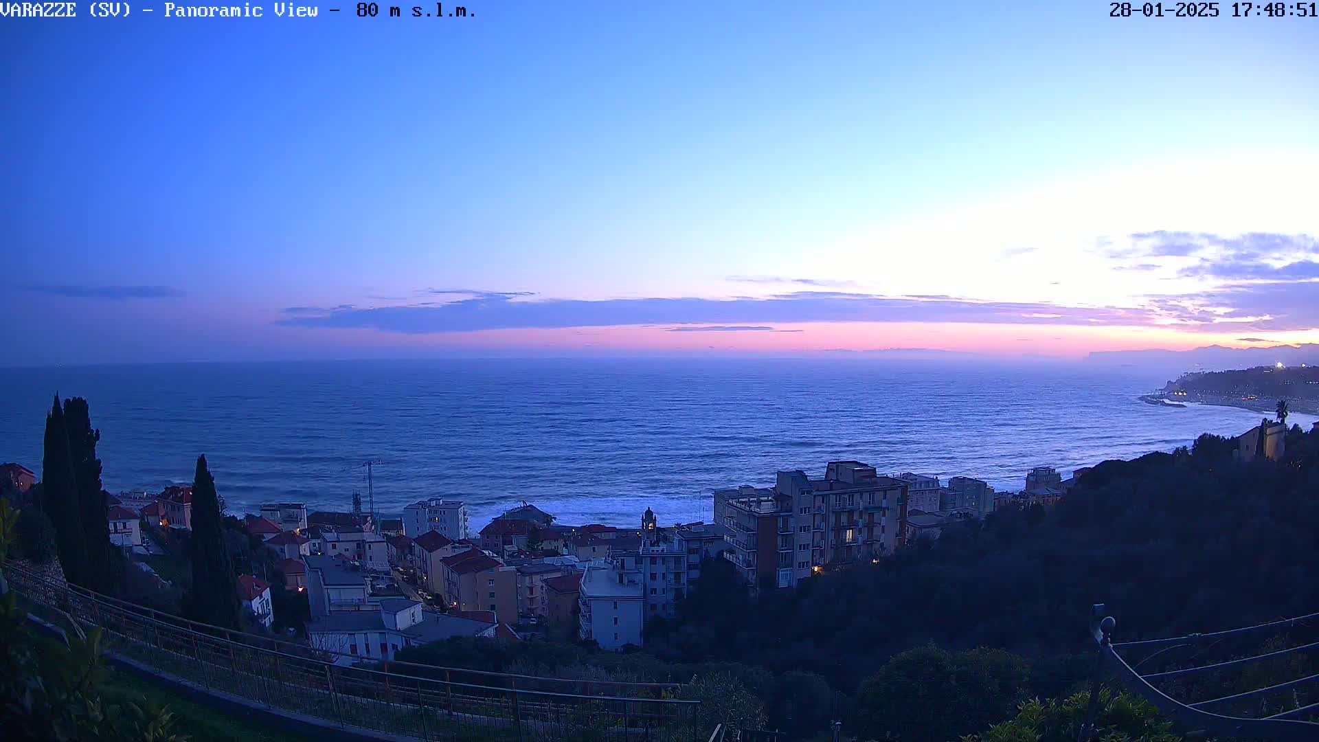 A coastal town at twilight overlooks a dark blue sea under a pale pink and purple sky.