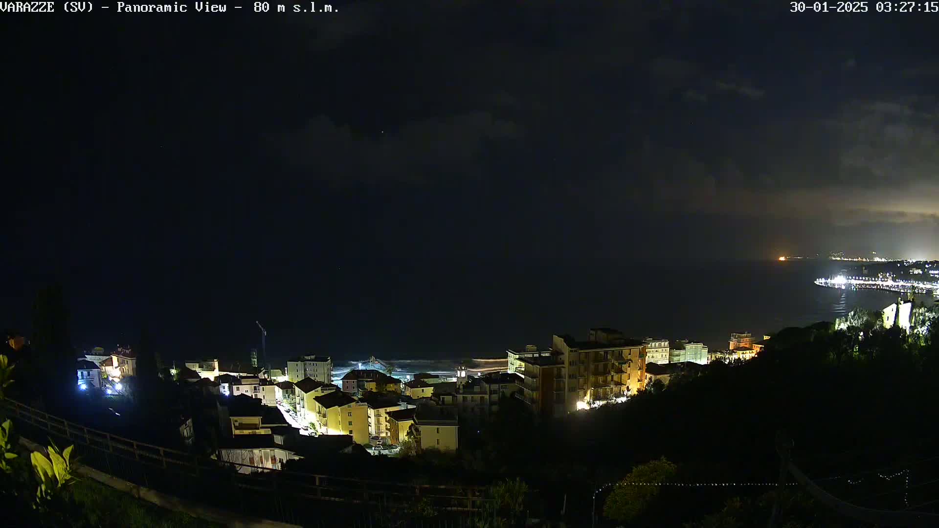 A nighttime, panoramic view of a coastal town with dimly lit buildings and a dark, cloudy sky over a calm sea.
