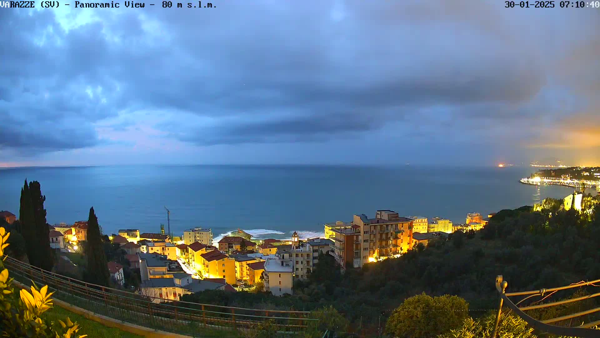 A coastal town with illuminated buildings is nestled on a hillside overlooking a calm sea under a cloudy twilight sky.