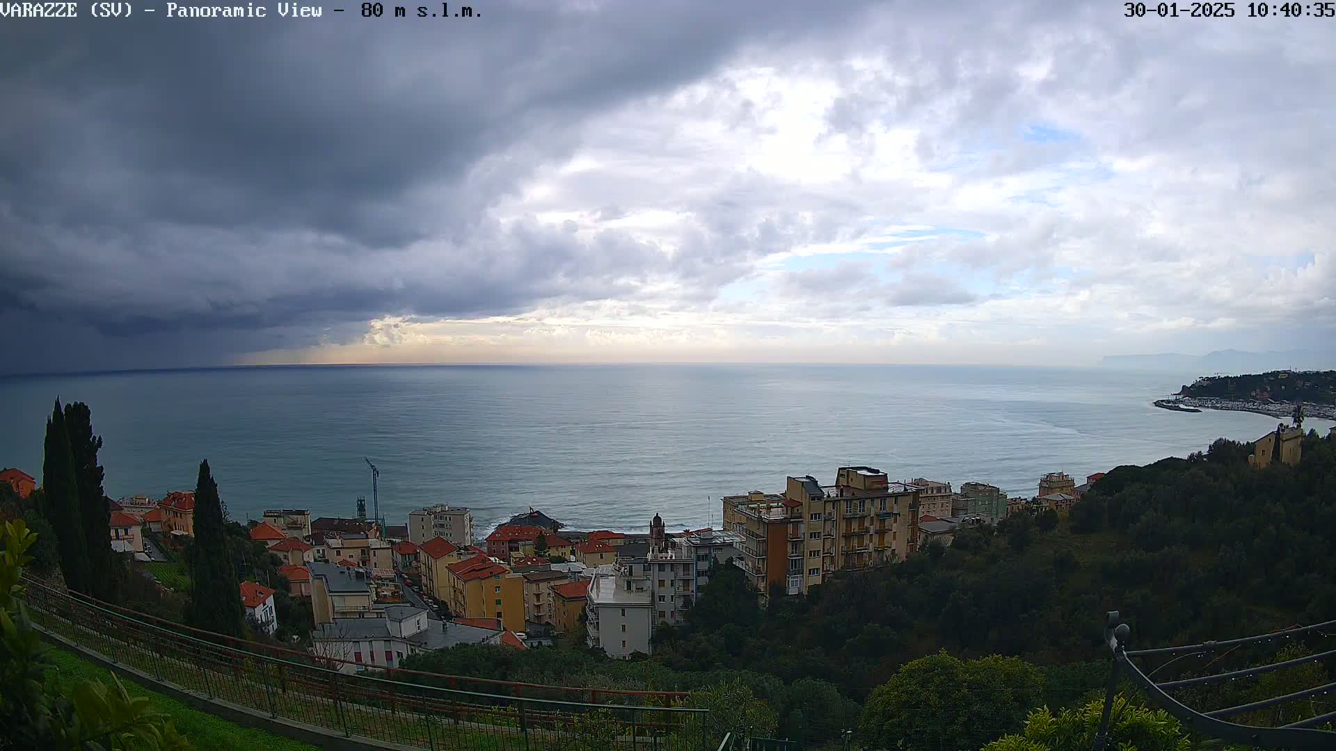 A panoramic view shows a coastal town nestled on a hillside overlooking a calm sea under a mostly cloudy sky with dark storm clouds in the distance.