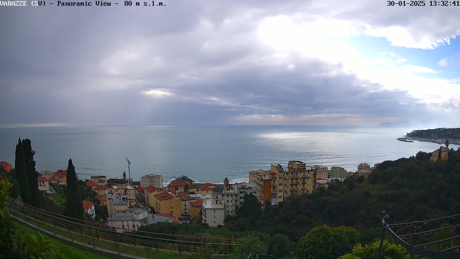 A coastal town with multi-story buildings sits on a hillside overlooking a calm sea under a mostly cloudy sky.