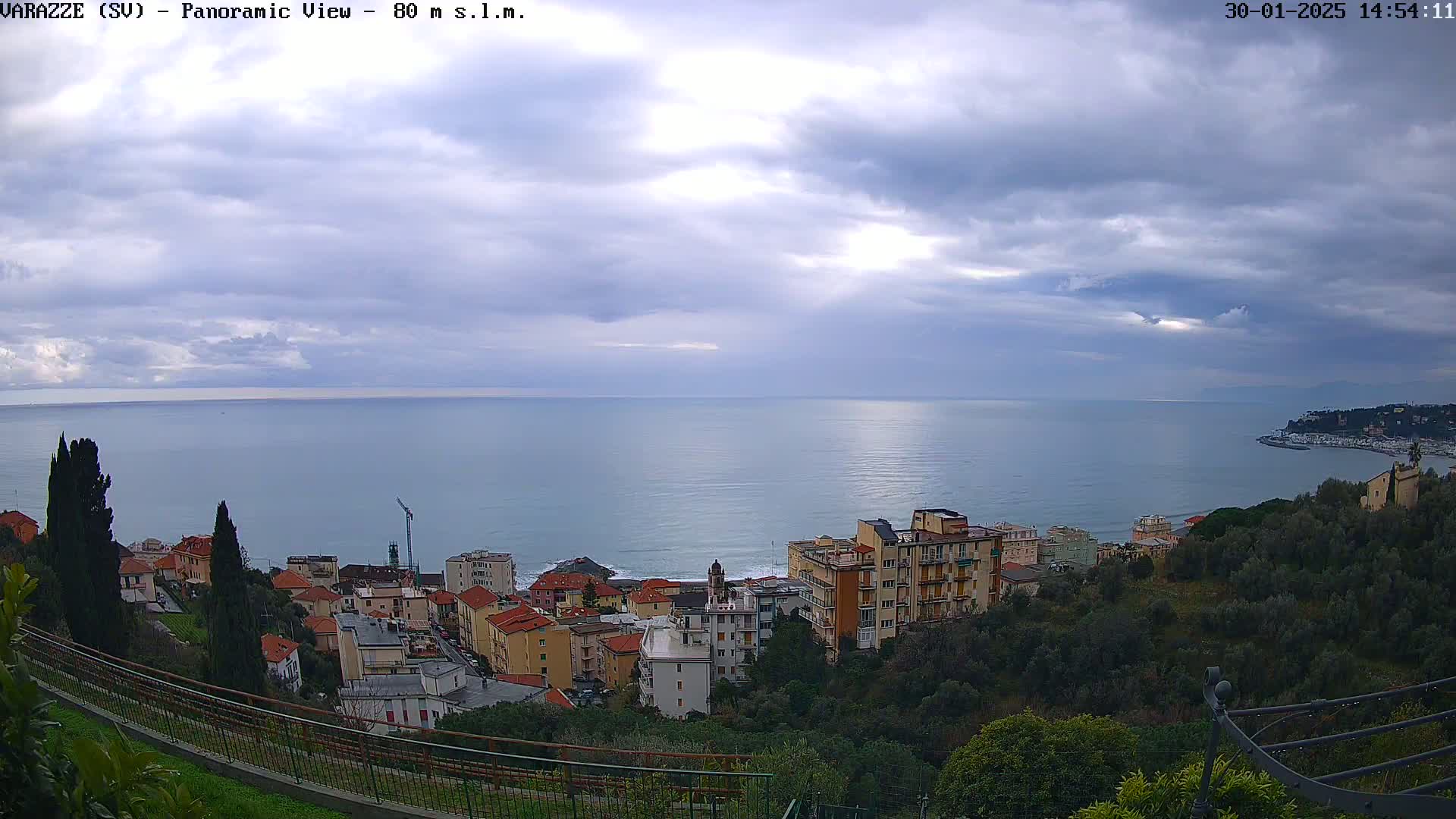 A coastal town with multi-story buildings is nestled on a hillside overlooking a calm sea under a mostly cloudy sky.