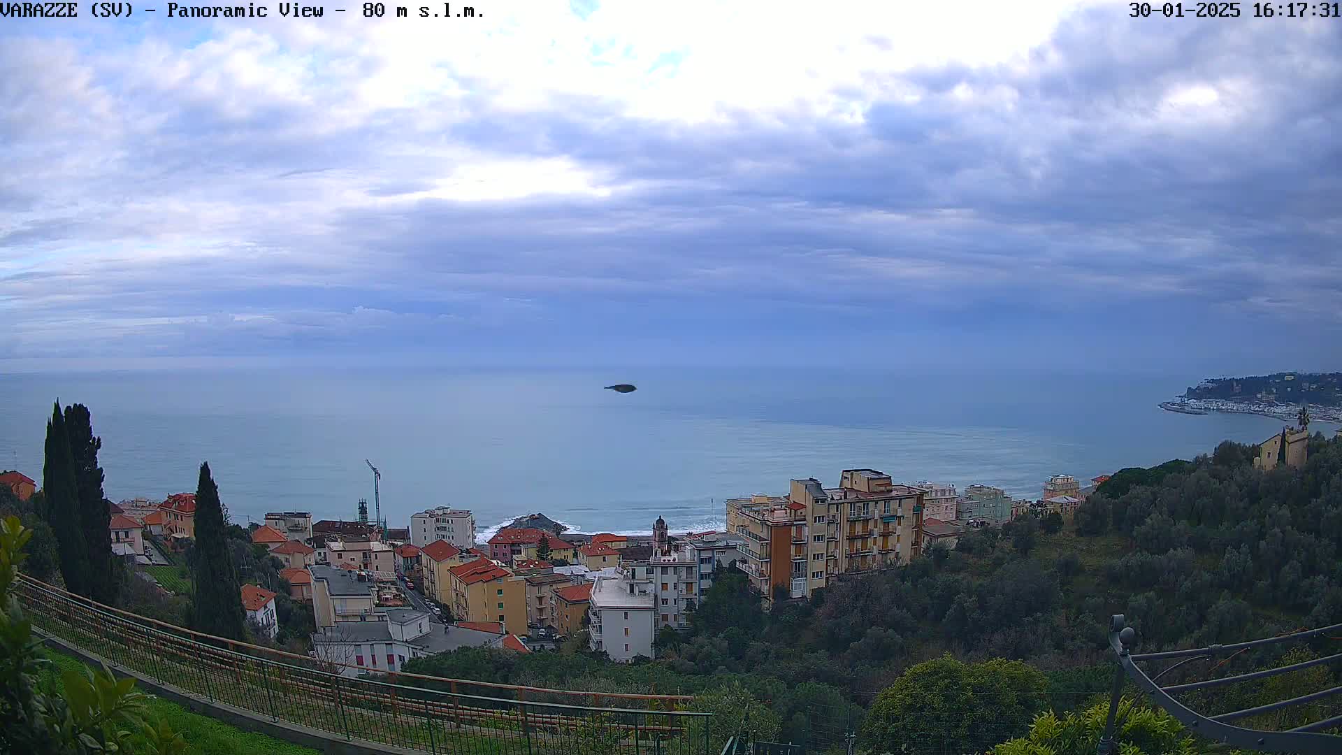 A coastal town with pastel-colored buildings sits on a hillside overlooking a calm sea under a mostly cloudy sky.