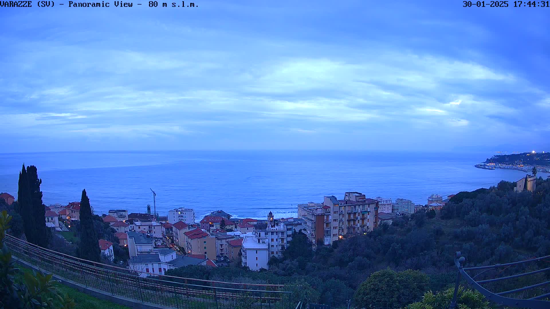 A coastal town nestled on a hillside overlooks a calm, blue sea under a twilight sky.