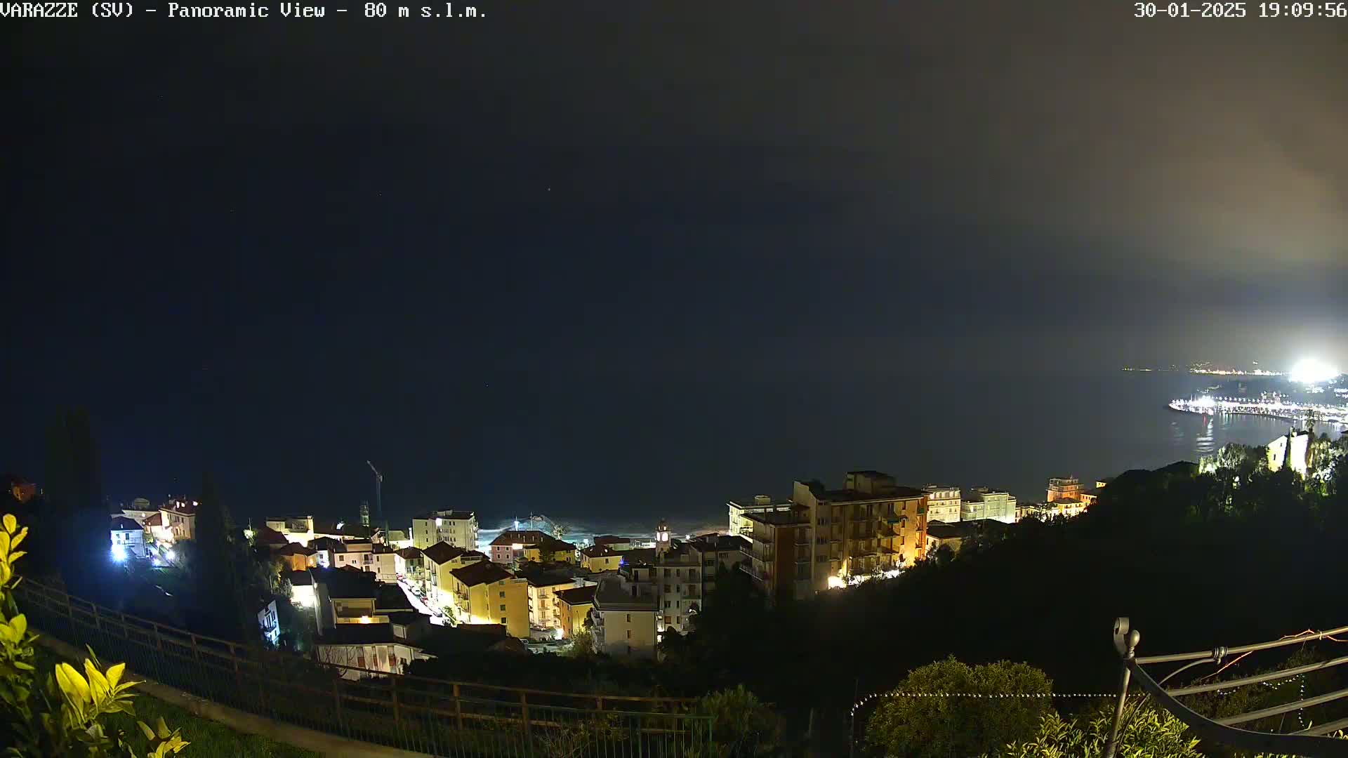 A nighttime panoramic view shows a coastal town illuminated, with a dark, calm sea extending to a dimly lit horizon under a mostly clear, dark sky.