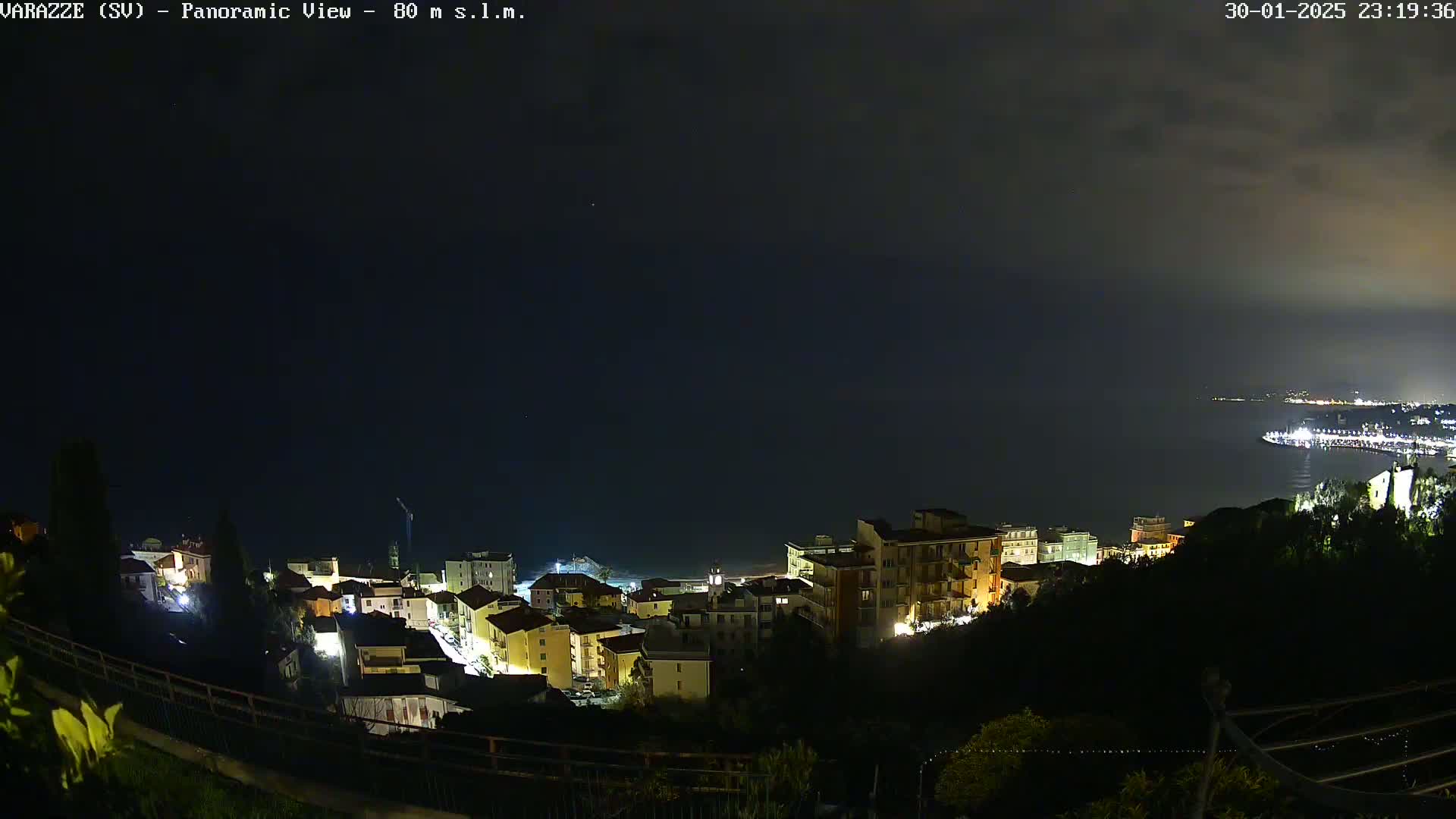 A nighttime panoramic view of a coastal town illuminated, with a dark, cloudy sky and a calm sea in the distance.