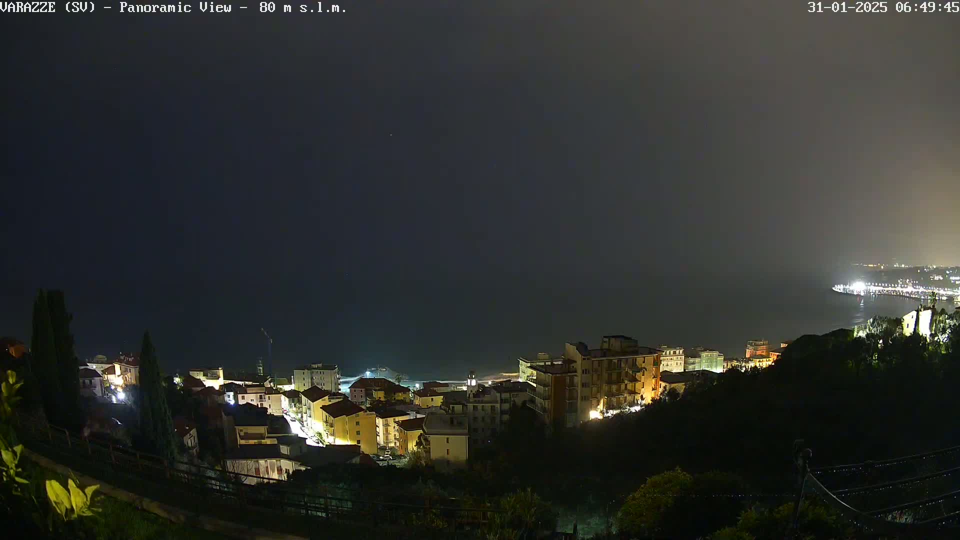 A nighttime panoramic view of a coastal town illuminated with lights, under a dark and overcast sky.