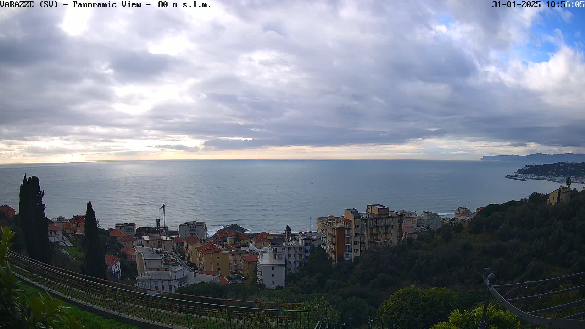 A panoramic view shows a coastal town nestled on a hillside overlooking a calm sea under a mostly cloudy sky.