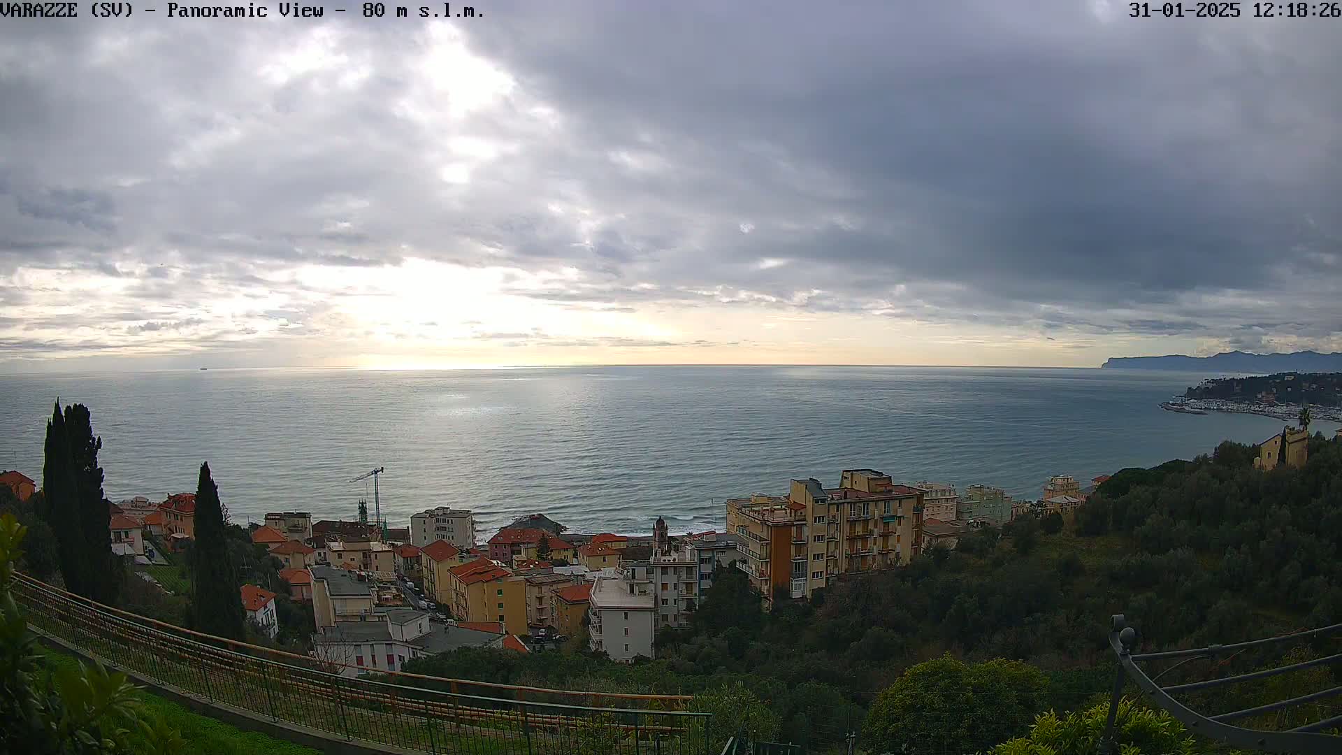A panoramic view shows a coastal town nestled on a hillside overlooking a calm sea under a cloudy sky.