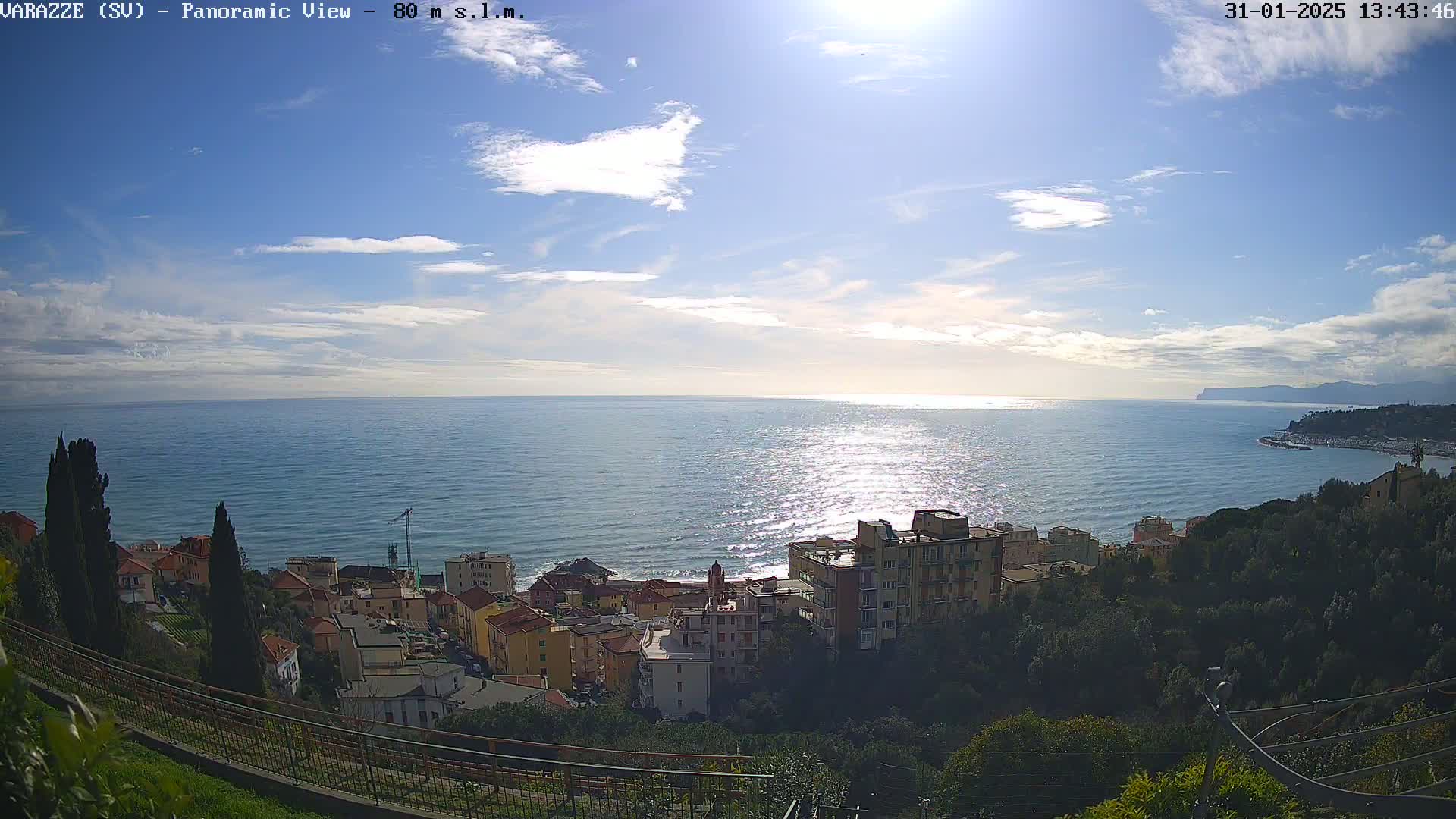 A panoramic view of a coastal town on a partly sunny day, with a calm sea stretching to the horizon under a mostly clear sky.