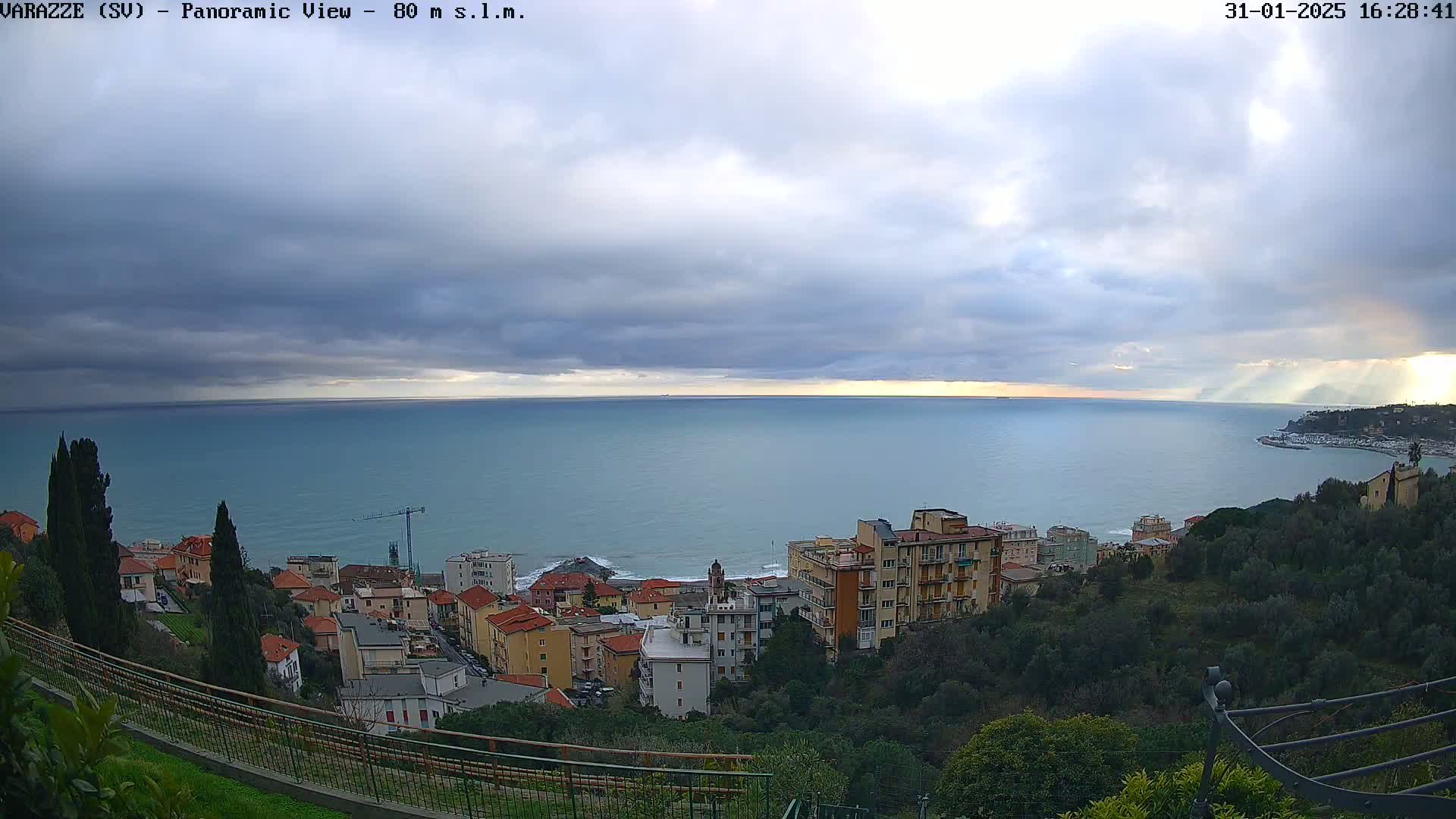 A panoramic view shows a coastal town nestled on a hillside overlooking a calm sea under a mostly cloudy sky with sun rays breaking through.