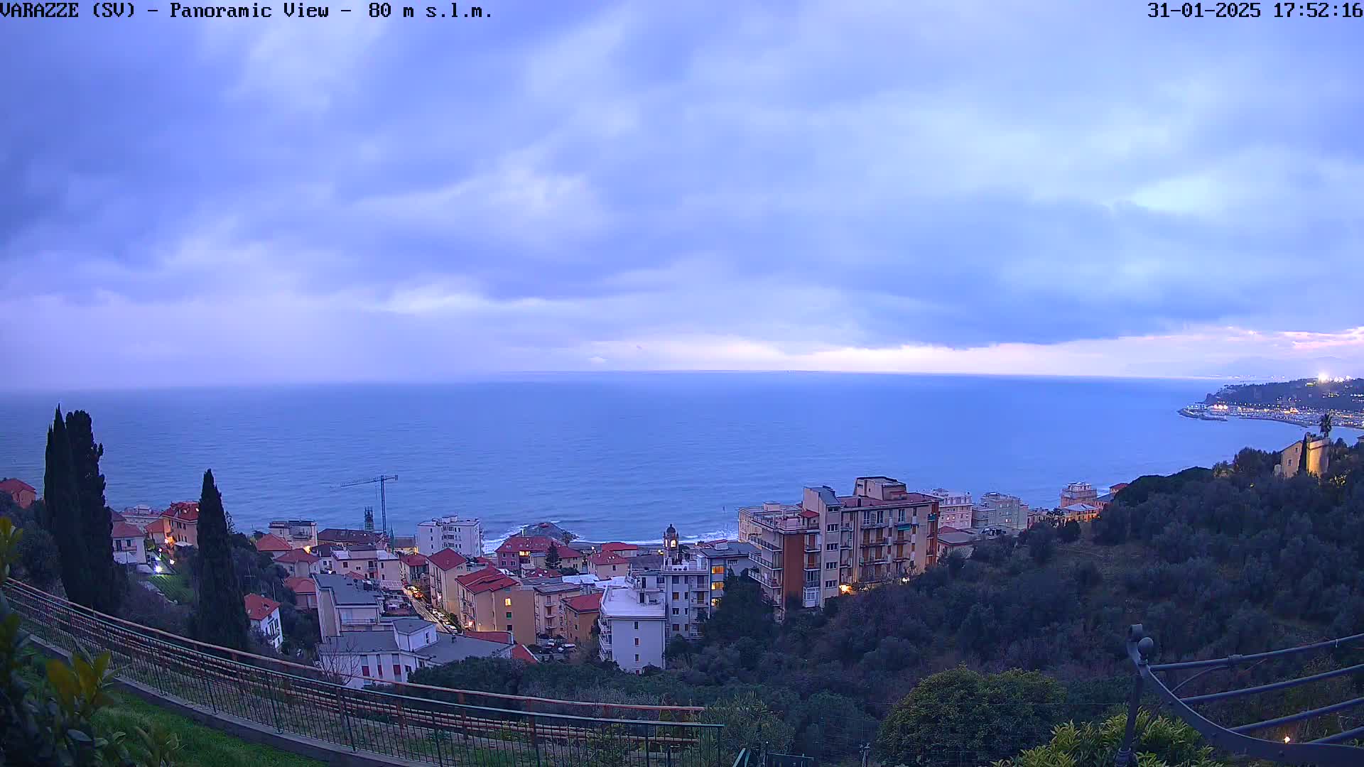 A coastal town overlooks a calm, blue sea under a cloudy, twilight sky.