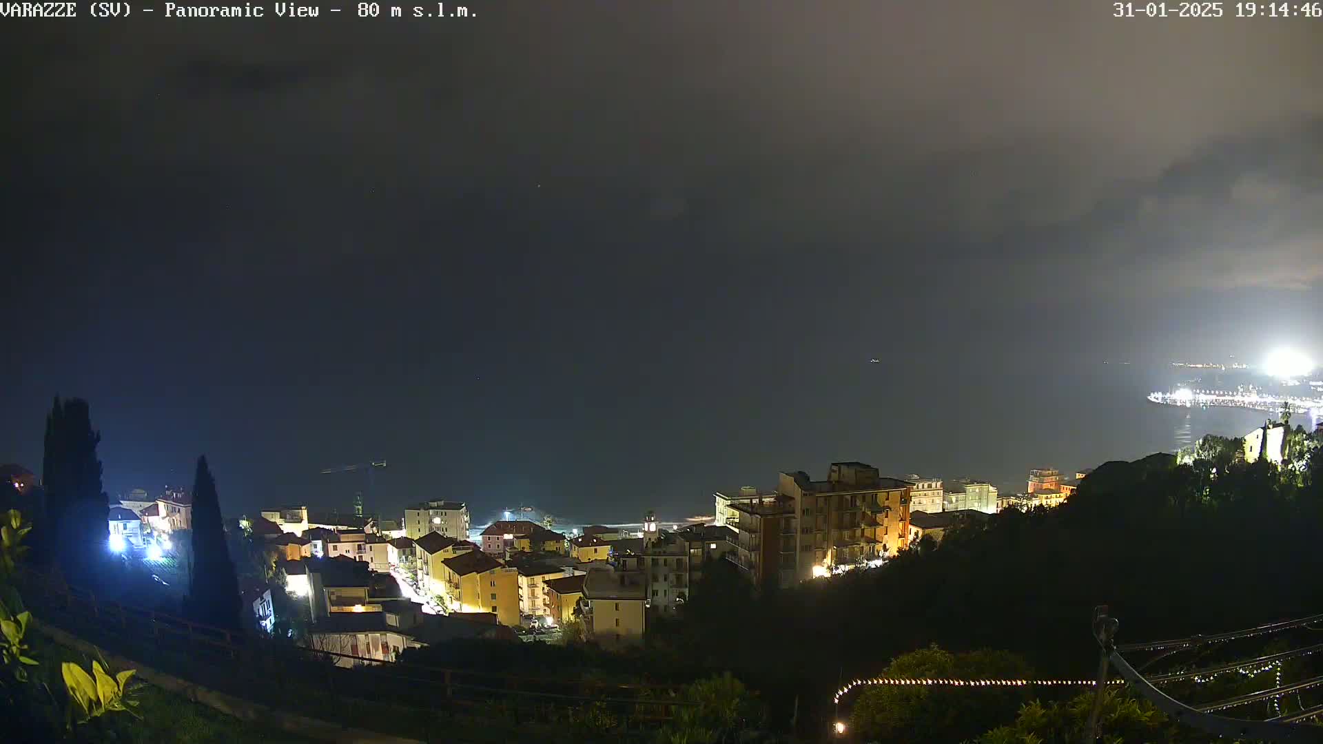 A nighttime panoramic view of a coastal town illuminated, with a dark, cloudy sky overhead and a calm sea in the distance.