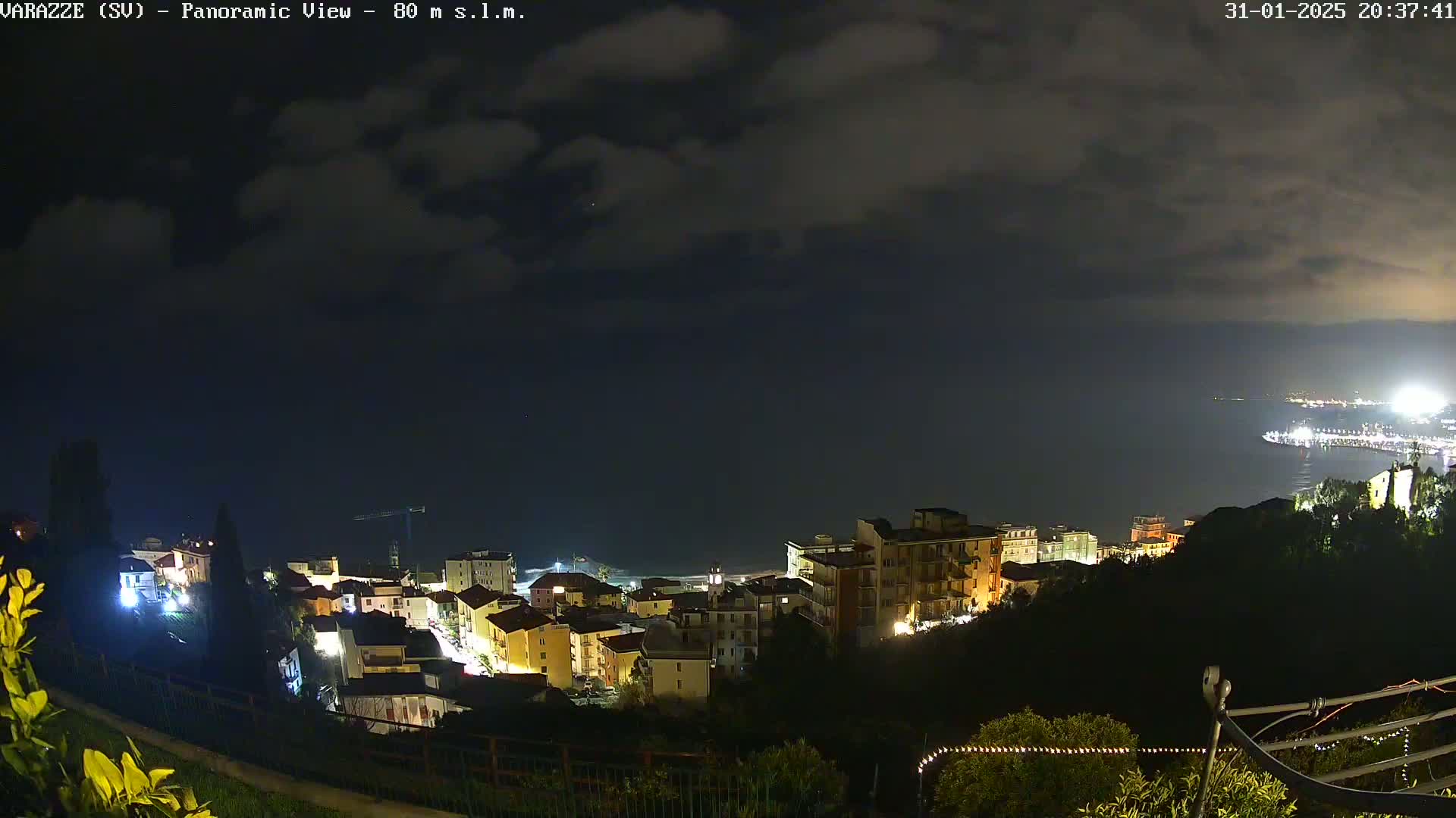 A nighttime panoramic view of a coastal town illuminated by lights, under a partly cloudy sky.