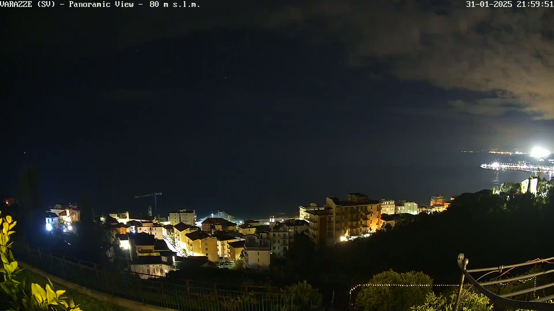 A nighttime panoramic view shows a coastal town illuminated with lights, under a partly cloudy sky, overlooking a dark body of water.