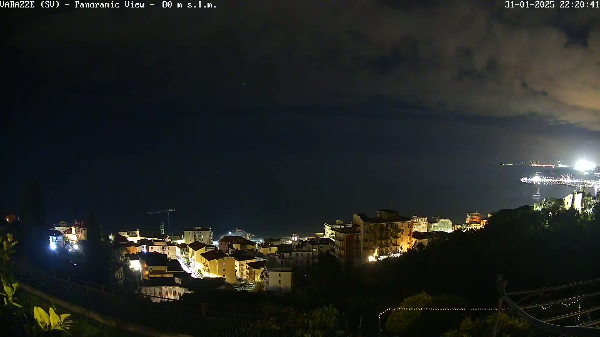 A nighttime panoramic view shows a coastal town illuminated under a partly cloudy sky.