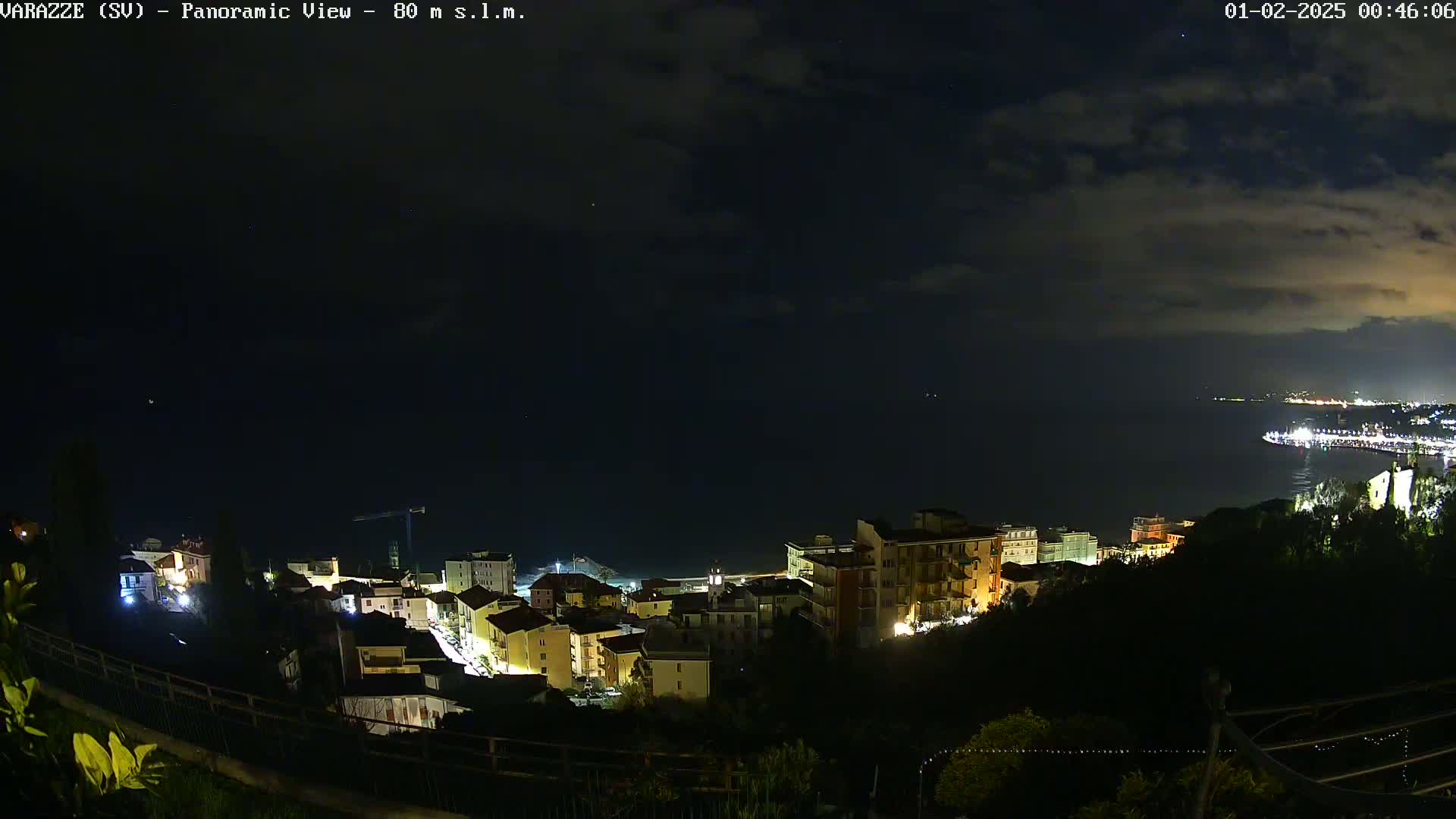 A nighttime panoramic view shows a coastal town with dimly lit buildings overlooking a dark sea under a partly cloudy sky.