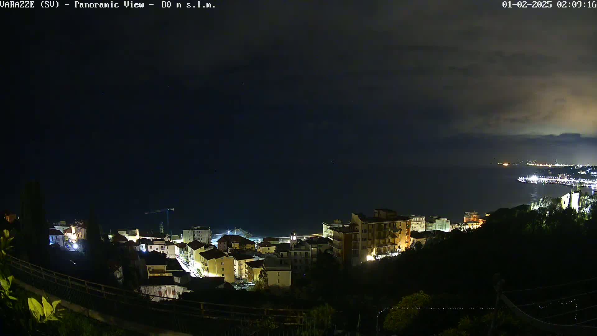 A nighttime, panoramic view of a coastal town with dimly lit buildings overlooking a dark ocean under a mostly cloudy sky.