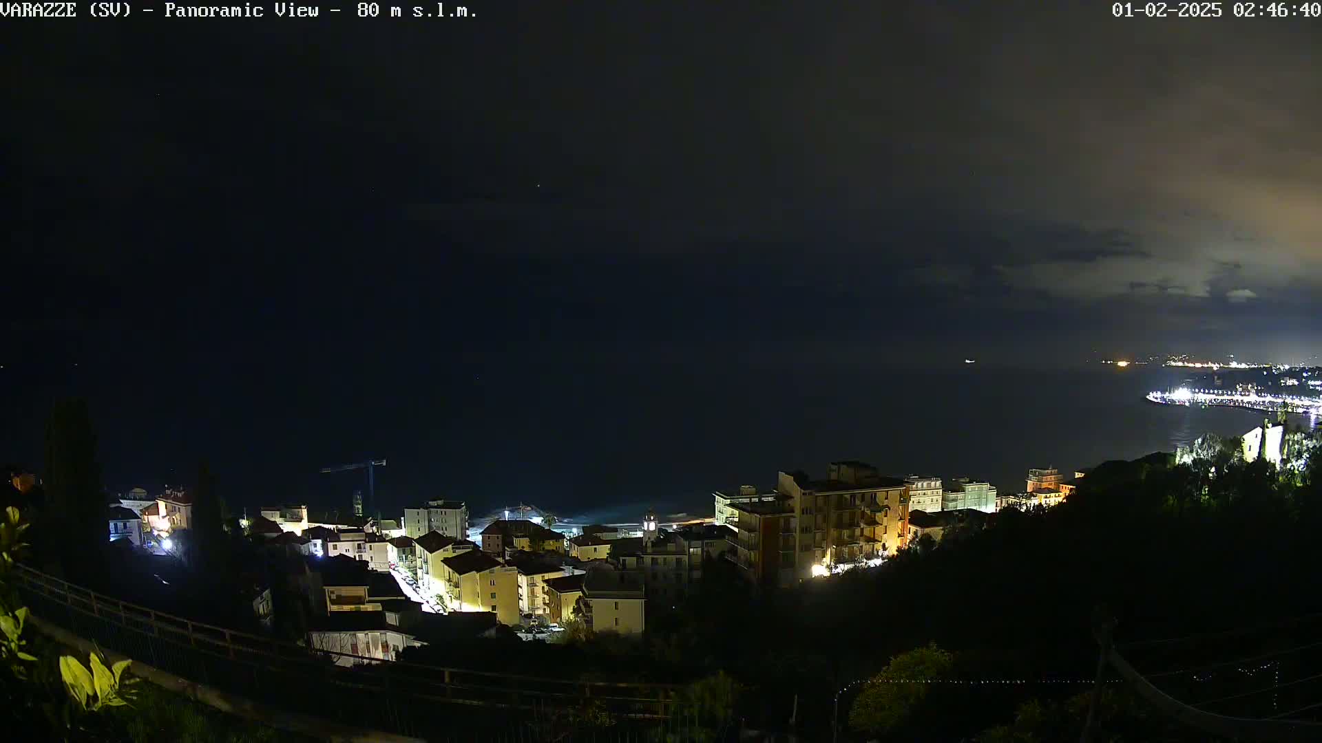 A nighttime panoramic view shows a coastal town with illuminated buildings, overlooking a dark sea under a mostly cloudy sky.