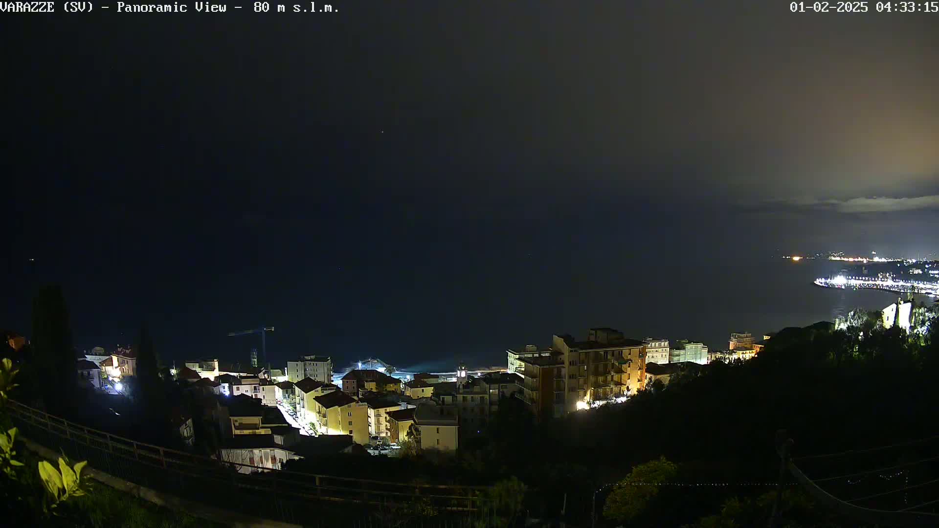 A nighttime, panoramic view of a coastal town with dimly lit buildings, overlooking a dark ocean under a cloudy sky.