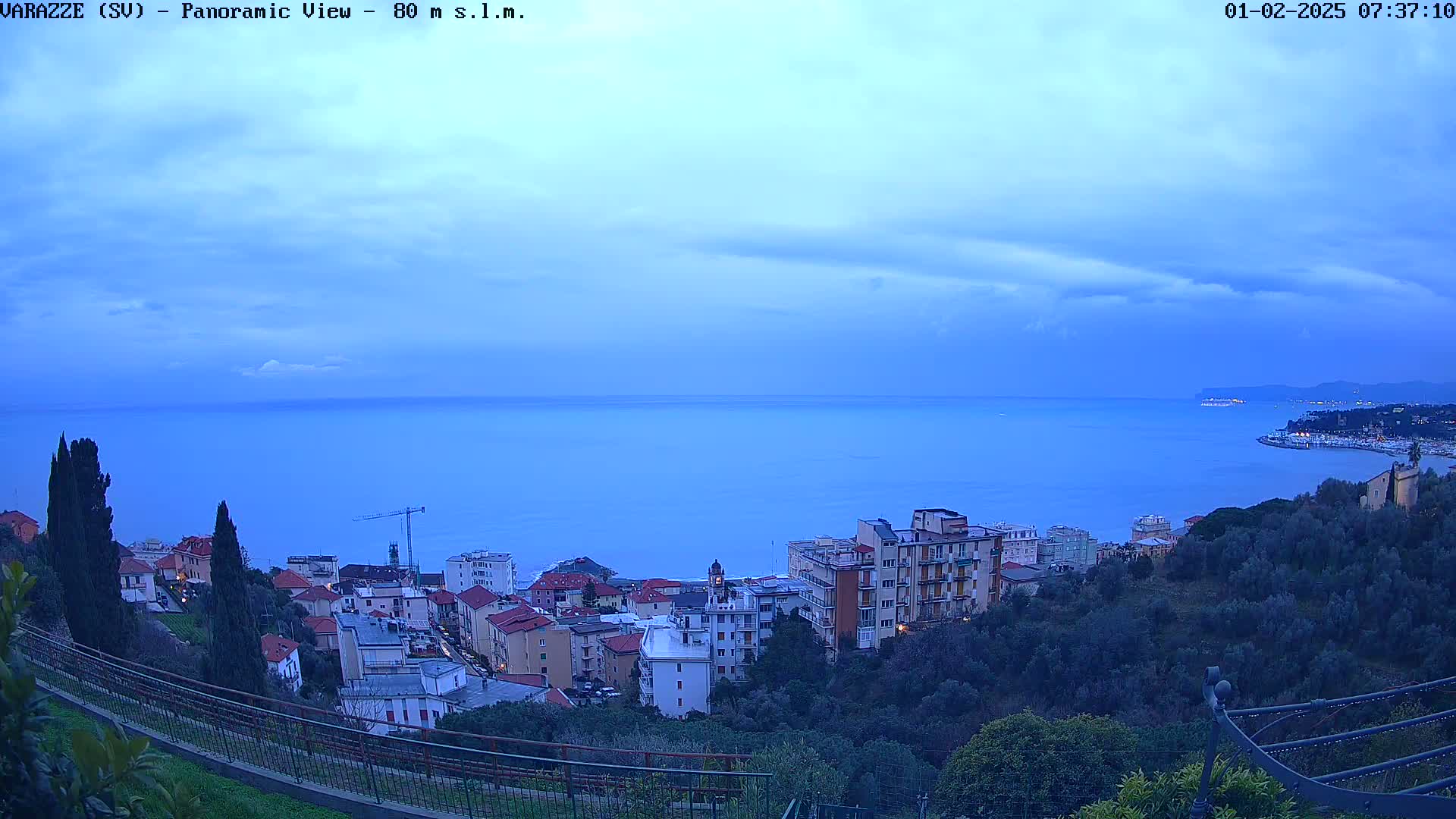 A panoramic view of a coastal town at twilight, under a mostly cloudy sky, shows a calm sea extending to a distant coastline.
