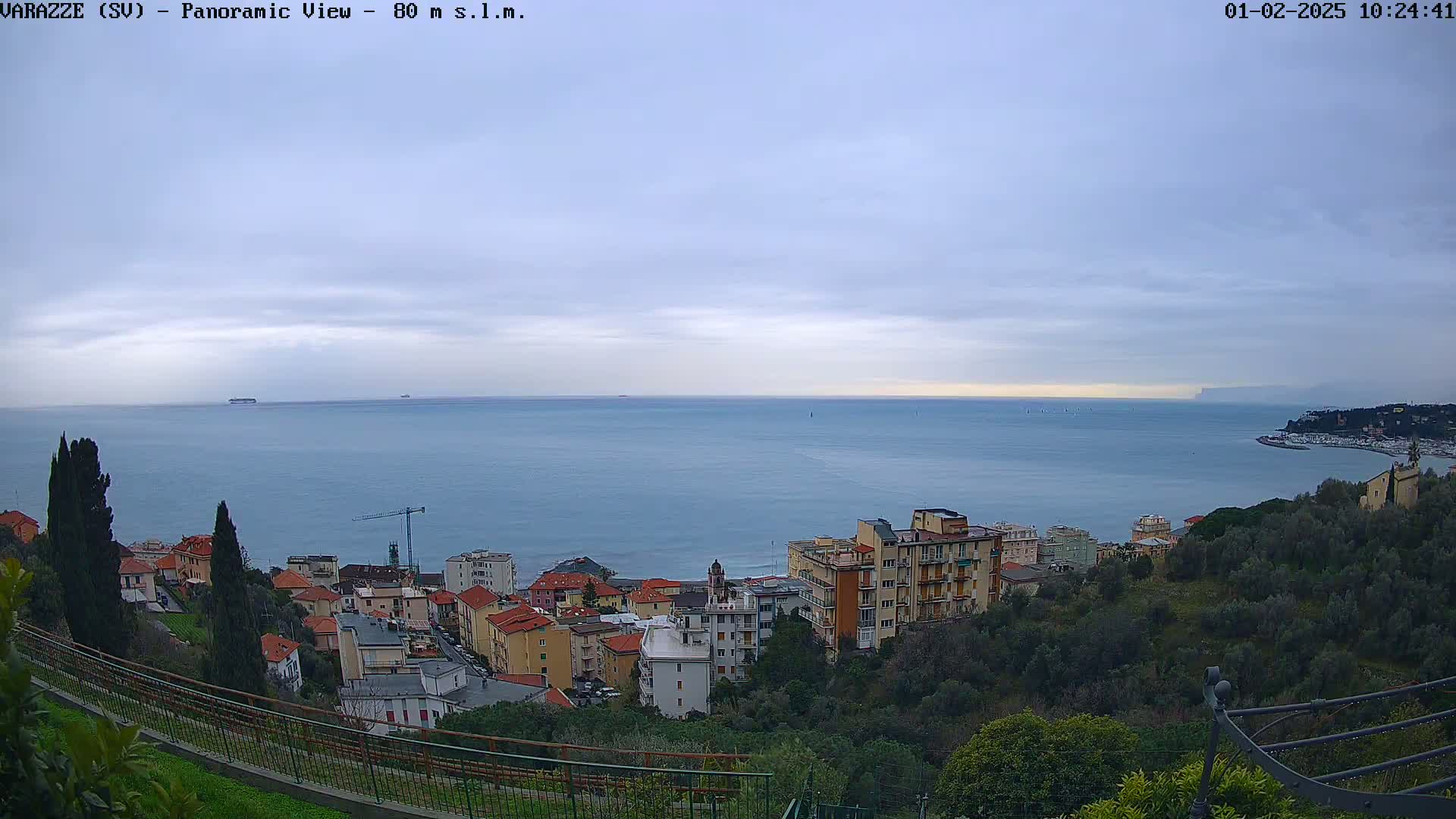 A panoramic view shows a coastal town nestled among green hills overlooking a calm, blue sea under a mostly cloudy sky.