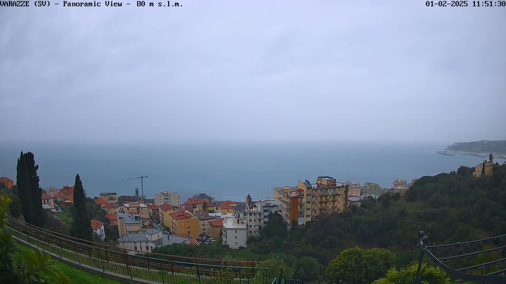 A panoramic view shows a coastal town on a hillside overlooking a calm sea under an overcast sky.