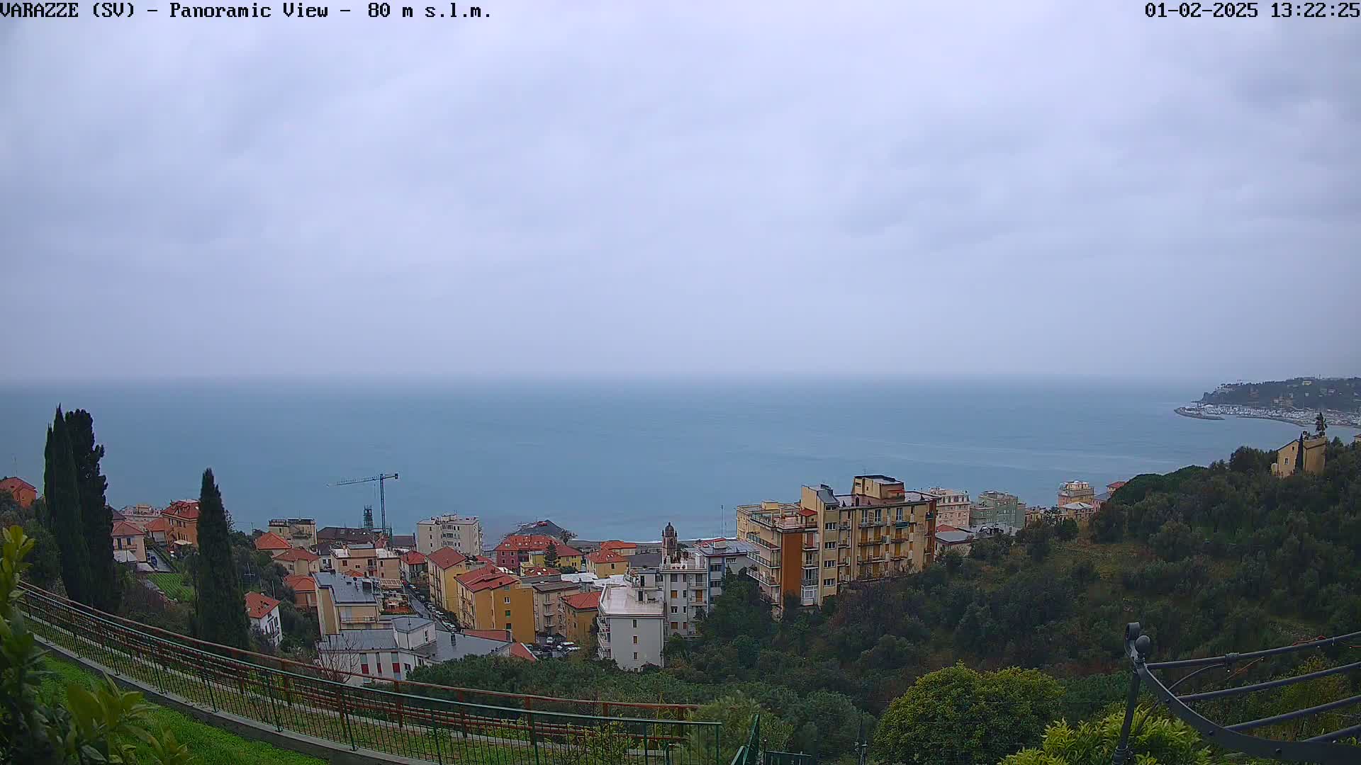 A panoramic view of a coastal town on a cloudy day, showing a calm sea and buildings nestled on a hillside.