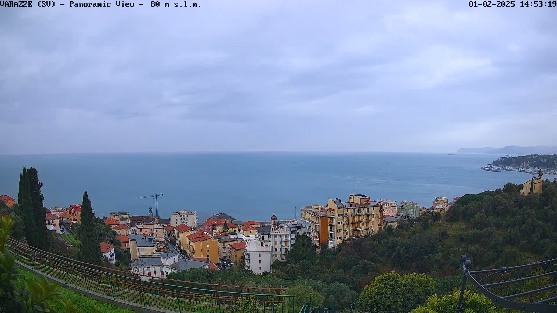 An overcast day reveals a coastal town nestled on a hillside overlooking a calm sea, with buildings of varying heights and colors spread across the landscape.