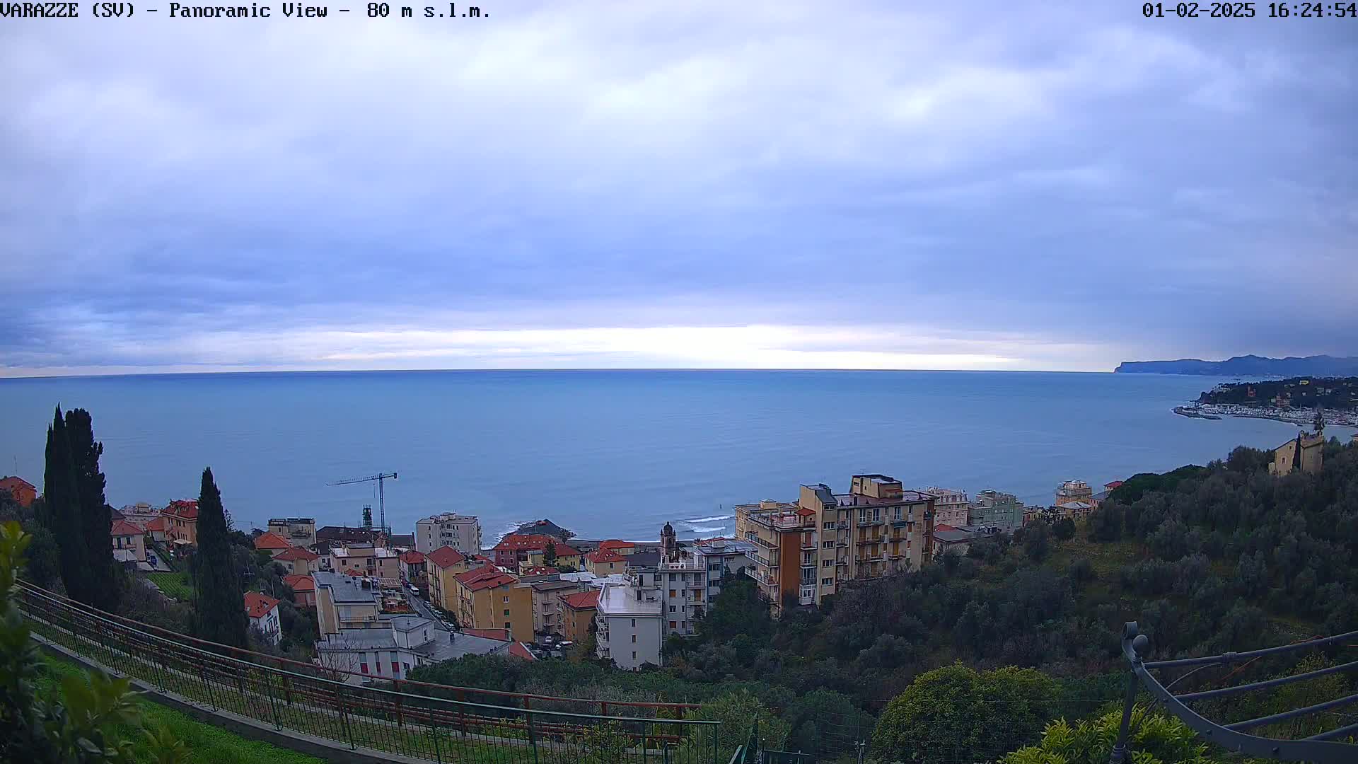 A panoramic view of a coastal town nestled on a hillside overlooking a calm sea under a mostly cloudy sky.