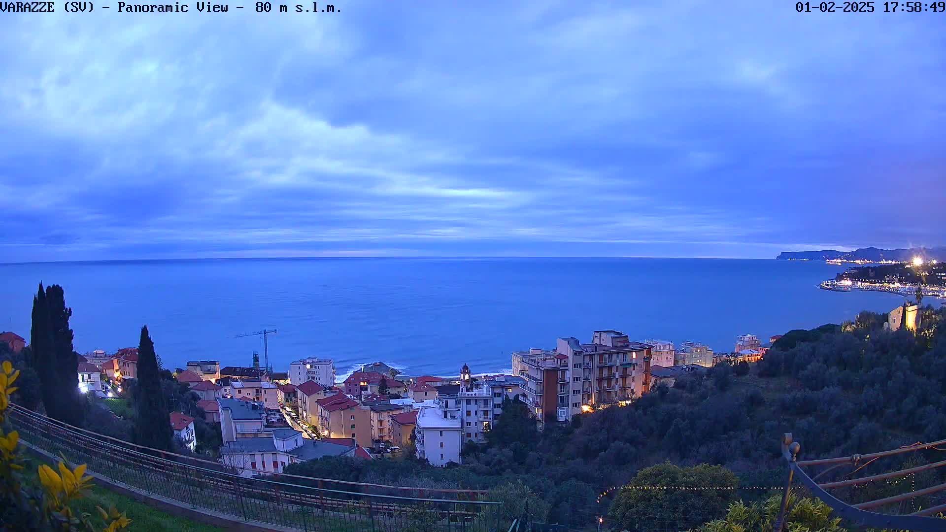 A coastal town at dusk is nestled on a hillside overlooking a calm, dark-blue sea under a cloudy twilight sky.