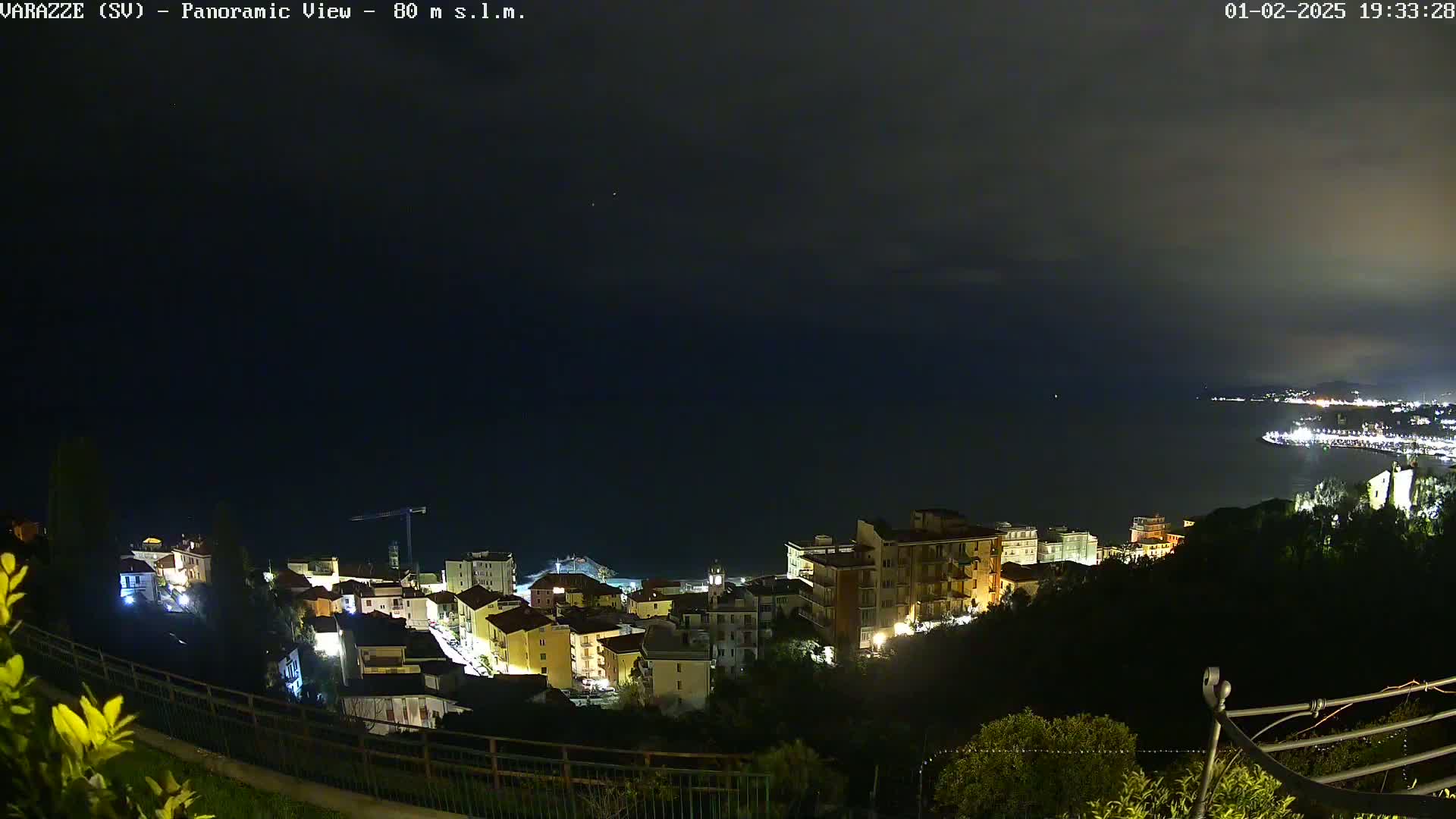 A nighttime panoramic view shows a coastal town with illuminated buildings overlooking a dark sea under a mostly cloudy sky.