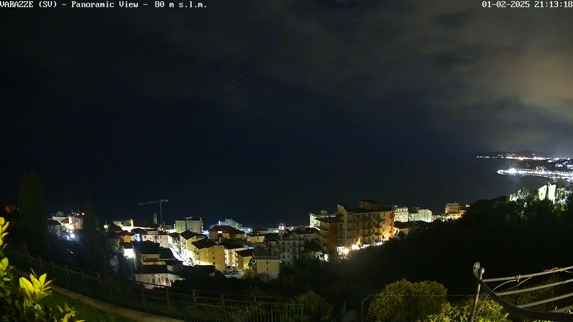 A nighttime panoramic view of a coastal town, illuminated with scattered lights, under a dark, cloudy sky.