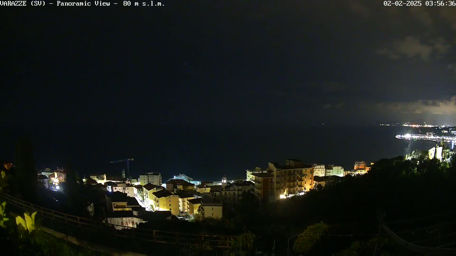 A nighttime panoramic view shows a coastal town with illuminated buildings nestled on a hillside overlooking a dark body of water under a mostly cloudy sky.
