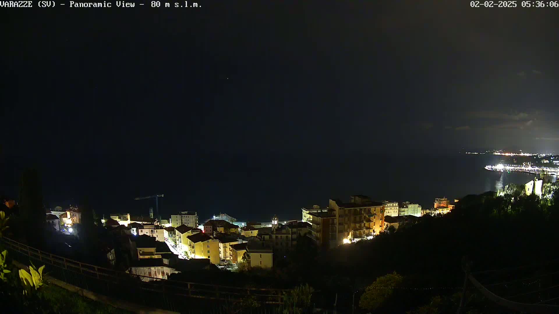A nighttime, panoramic view of a coastal town with its buildings illuminated, overlooking a dark sea under a clear sky.