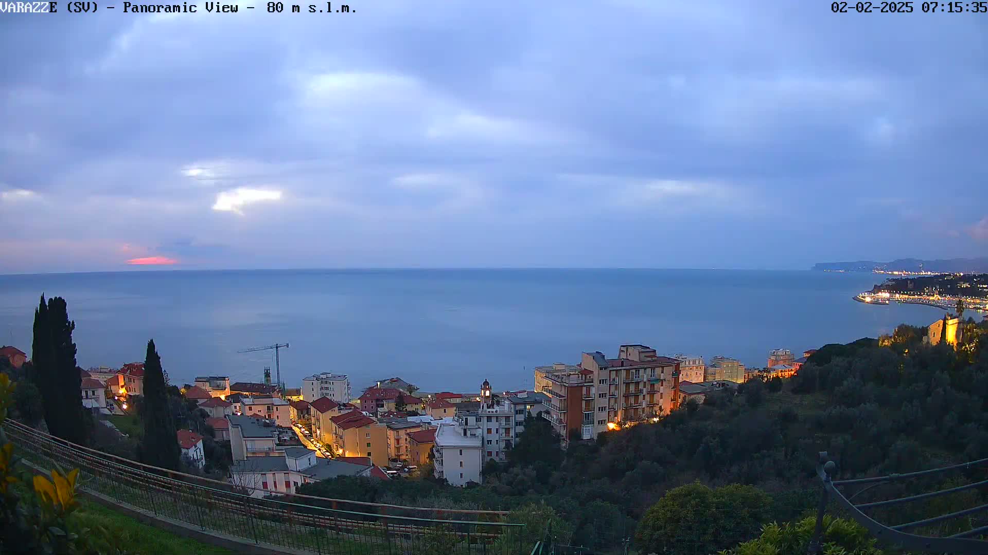 A coastal town at twilight is nestled on a hillside overlooking a calm sea under a mostly cloudy sky.