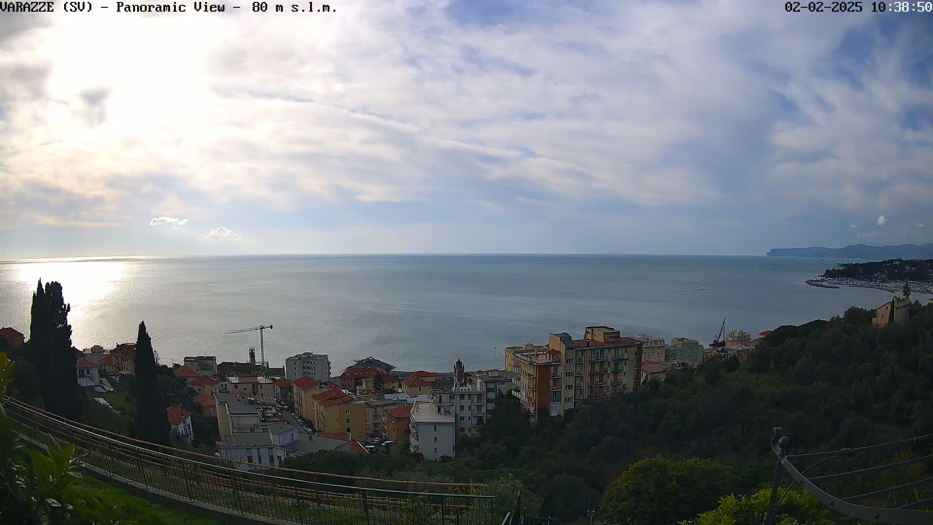 A panoramic view of a coastal town nestled on a hillside overlooking a calm sea under a partly cloudy sky.