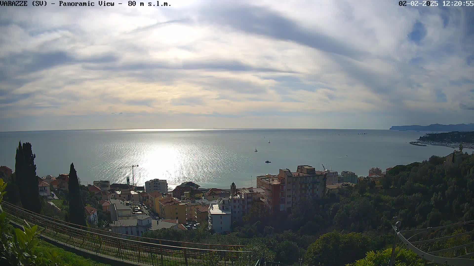 A panoramic view of a coastal town under a partly cloudy sky shows a calm sea reflecting sunlight, with boats and buildings visible in the distance.