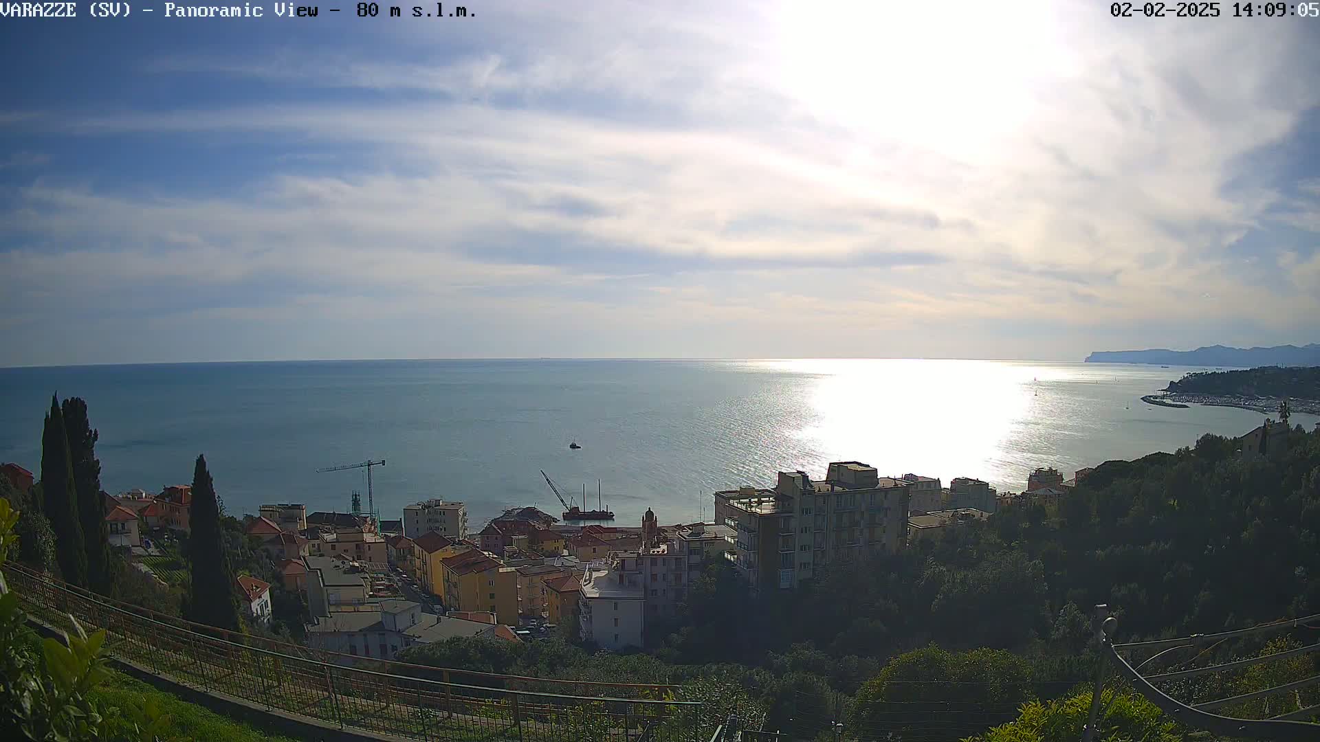 A panoramic view of a coastal town nestled on a hillside overlooking a calm, sunlit sea under a partly cloudy sky.