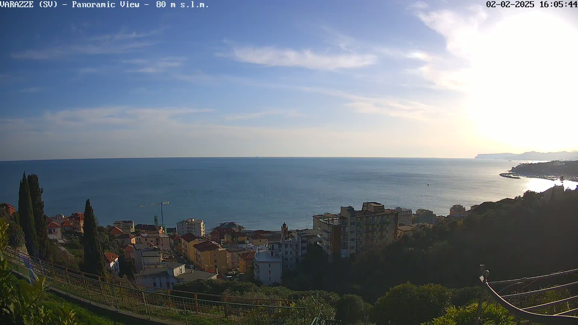 A coastal town with multi-story buildings sits on a hillside overlooking a calm sea under a partly cloudy sky.