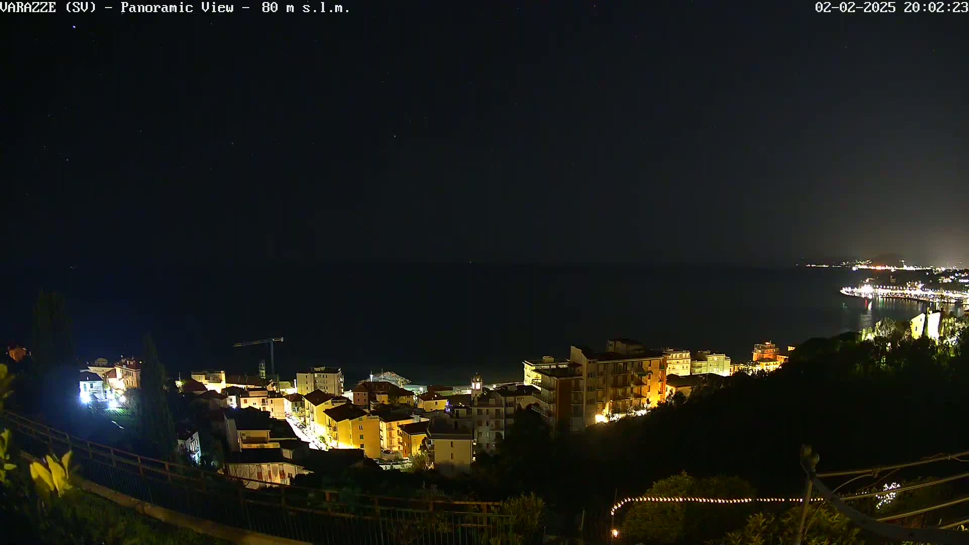 A nighttime panoramic view of a coastal town illuminated with lights, overlooking a dark sea under a clear, starry sky.