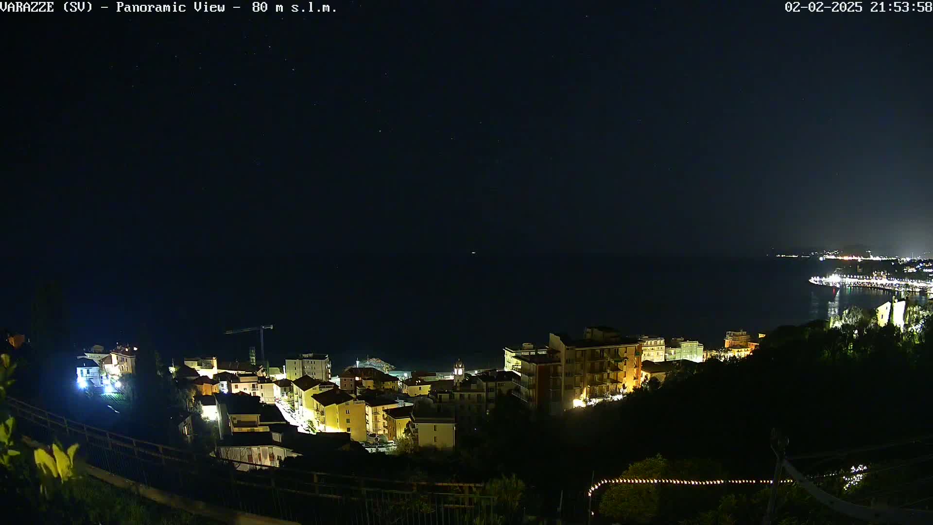 A nighttime panoramic view shows a coastal town with illuminated buildings overlooking a dark sea under a clear, starry sky.