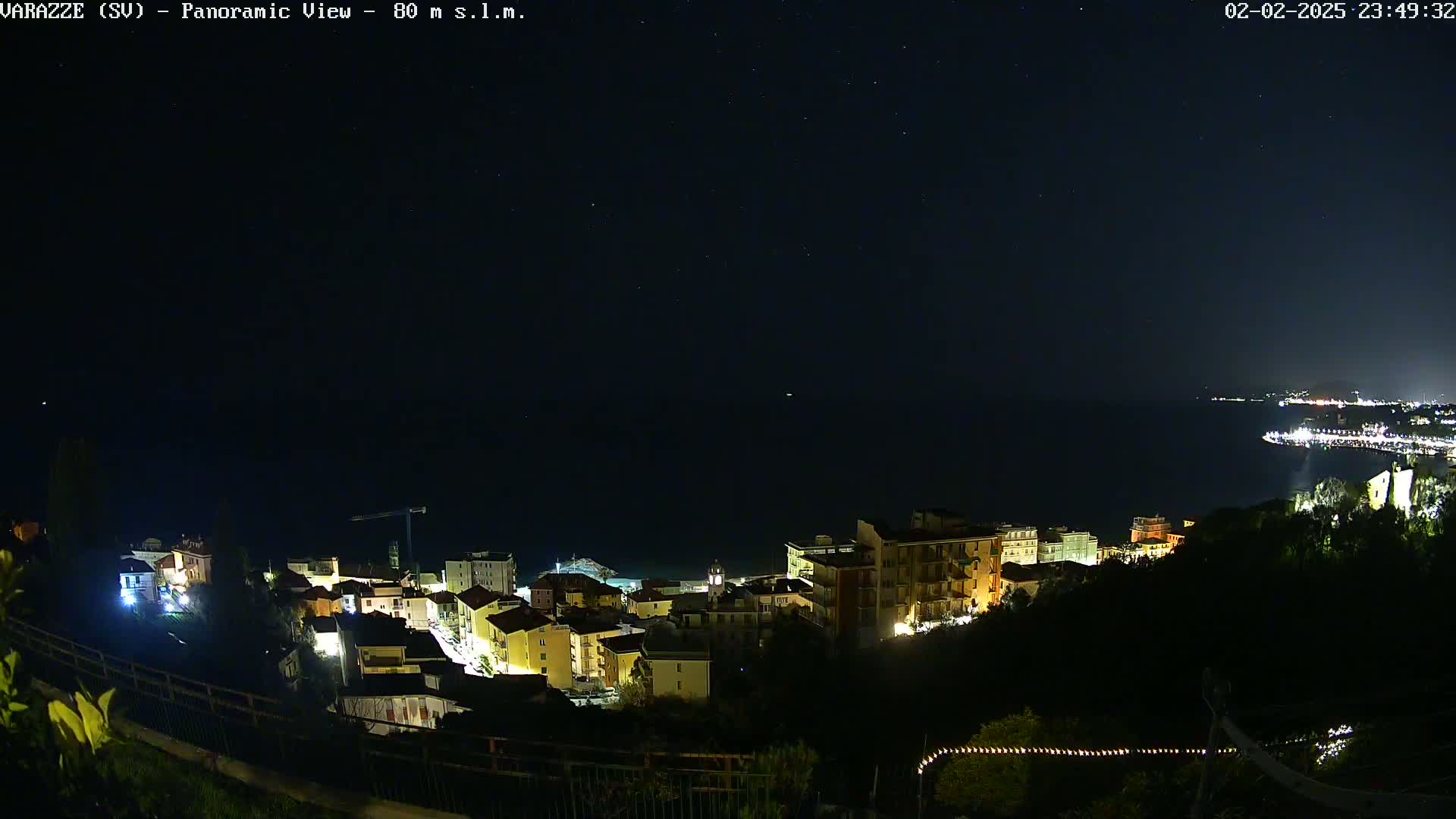 A nighttime panoramic view shows a coastal town illuminated, with a dark sea and starry sky above.
