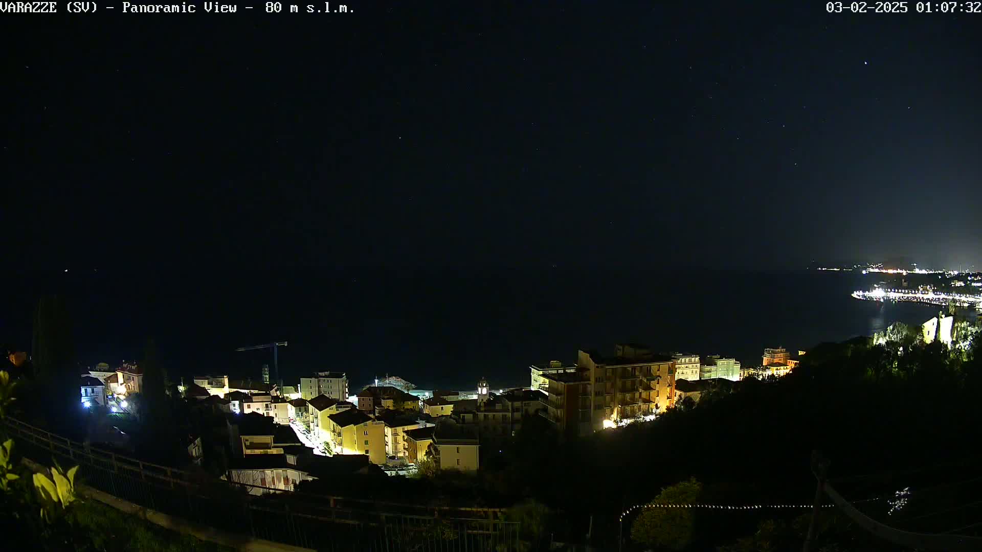 A nighttime panoramic view of a coastal town with its lights illuminated against a dark, clear sky speckled with stars.